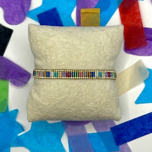 May include: A colourful beaded bracelet with a gold accent on a white cushion. The bracelet is made of small, colourful beads and has a gold metal clasp.