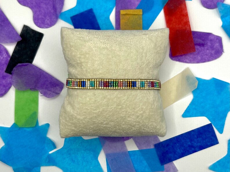 May include: A colourful beaded bracelet with a gold accent on a white cushion. The bracelet is made of small, colourful beads and has a gold metal clasp.