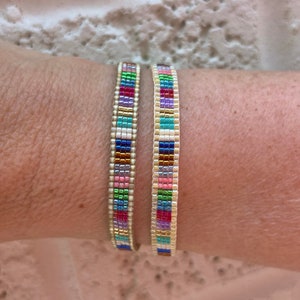 May include: Two colourful beaded bracelets with a gold thread running through the centre of each bracelet. The bracelets are on a person's wrist.
