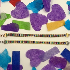 May include: Two beaded bracelets with colourful beads and silver and gold clasps. The bracelets are on a white surface with colourful tissue paper hearts and stars.