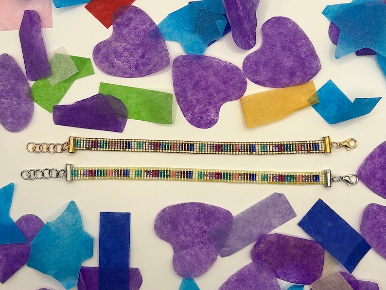 May include: Two beaded bracelets with colourful beads and silver and gold clasps. The bracelets are on a white surface with colourful tissue paper hearts and stars.