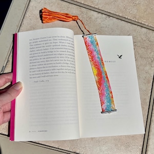 May include: A colorful beaded bookmark with a tassel is placed in an open book. The bookmark features a vibrant pattern of red, yellow, and blue beads. The book's pages are white, and the spine is pink. The word "FOREWORD" is visible.