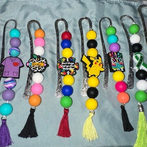 May include: Ten colorful beaded bookmarks with metal clips and tassels. Each bookmark features a different charm, including a pink ribbon, a nurse's uniform, an apple with the word "Teacher", a puzzle piece with the text "Don't Judge What You Don't Understand", a Pikachu, a box of crayons, a green ribbon with the text "Never Give Up", and a cartoon orange character.