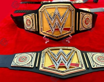 Puede incluir: Dos cinturones de campeonato dorados y negros con el logo de la WWE. Los cinturones tienen placas centrales doradas con el logo de la WWE y las palabras "Undisputed Champion". Los cinturones están sobre una superficie roja.