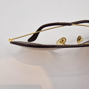 K&ouml;nnte beinhalten: Nahaufnahme einer Sonnenbrille mit einem braunen, strukturierten Rahmen und goldfarbenen Metallb&uuml;geln. Die Gl&auml;ser sind dunkel und das Gesamtdesign ist elegant und klassisch. Die Brille ist vor einem wei&szlig;en Hintergrund platziert.