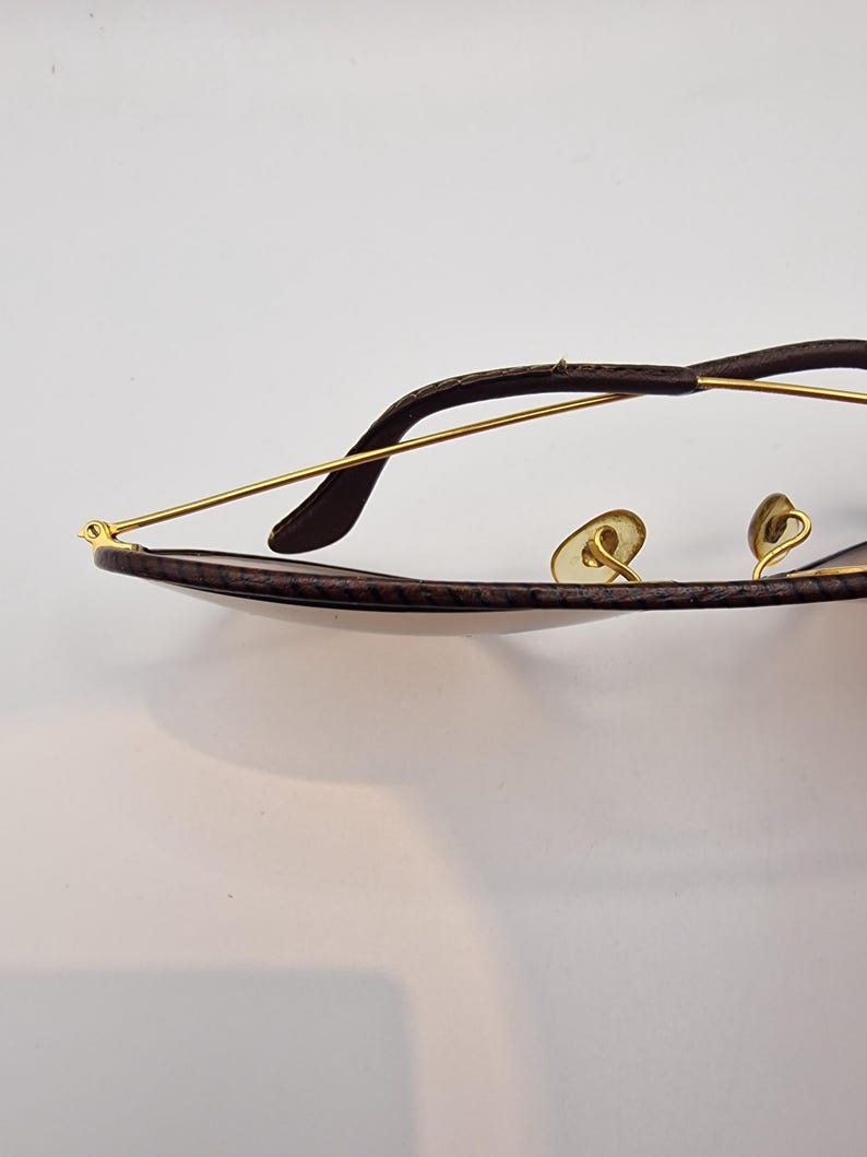 K&ouml;nnte beinhalten: Nahaufnahme einer Sonnenbrille mit einem braunen, strukturierten Rahmen und goldfarbenen Metallb&uuml;geln. Die Gl&auml;ser sind dunkel und das Gesamtdesign ist elegant und klassisch. Die Brille ist vor einem wei&szlig;en Hintergrund platziert.