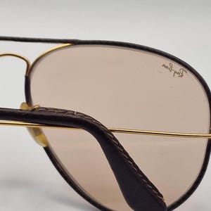 K&ouml;nnte beinhalten: Nahaufnahme einer Pilotenbrille. Der braune Lederb&uuml;gel und der goldfarbene Metallrahmen kontrastieren mit den hellbraunen Gl&auml;sern. Das Ray-Ban-Logo ist auf dem Glas zu sehen.