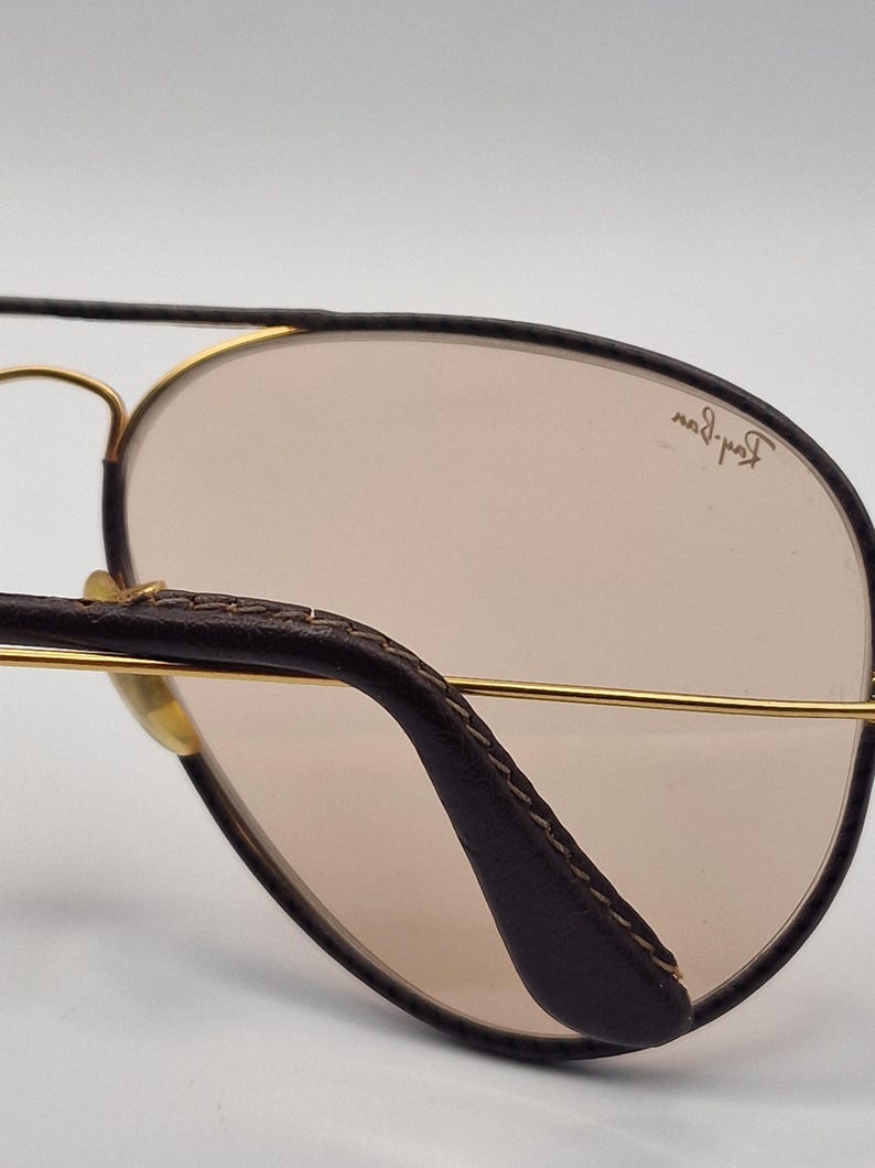K&ouml;nnte beinhalten: Nahaufnahme einer Pilotenbrille. Der braune Lederb&uuml;gel und der goldfarbene Metallrahmen kontrastieren mit den hellbraunen Gl&auml;sern. Das Ray-Ban-Logo ist auf dem Glas zu sehen.