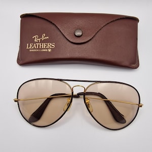 K&ouml;nnte beinhalten: Aviator-Sonnenbrille mit goldfarbenem Gestell und braunen Gl&auml;sern, zusammen mit einem braunen Lederetui. Das Etui ist mit dem Ray-Ban Leathers-Logo gepr&auml;gt. Die Sonnenbrille hat ein klassisches Design.