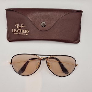K&ouml;nnte beinhalten: Braunes Leder-Sonnenbrillenetui mit dem Text "Ray-Ban LEATHERS BAUSCH & LOMB" und einer Pilotenbrille. Die Sonnenbrille hat goldfarbene Rahmen und braune Gl&auml;ser. Das Etui hat einen Druckknopfverschluss.