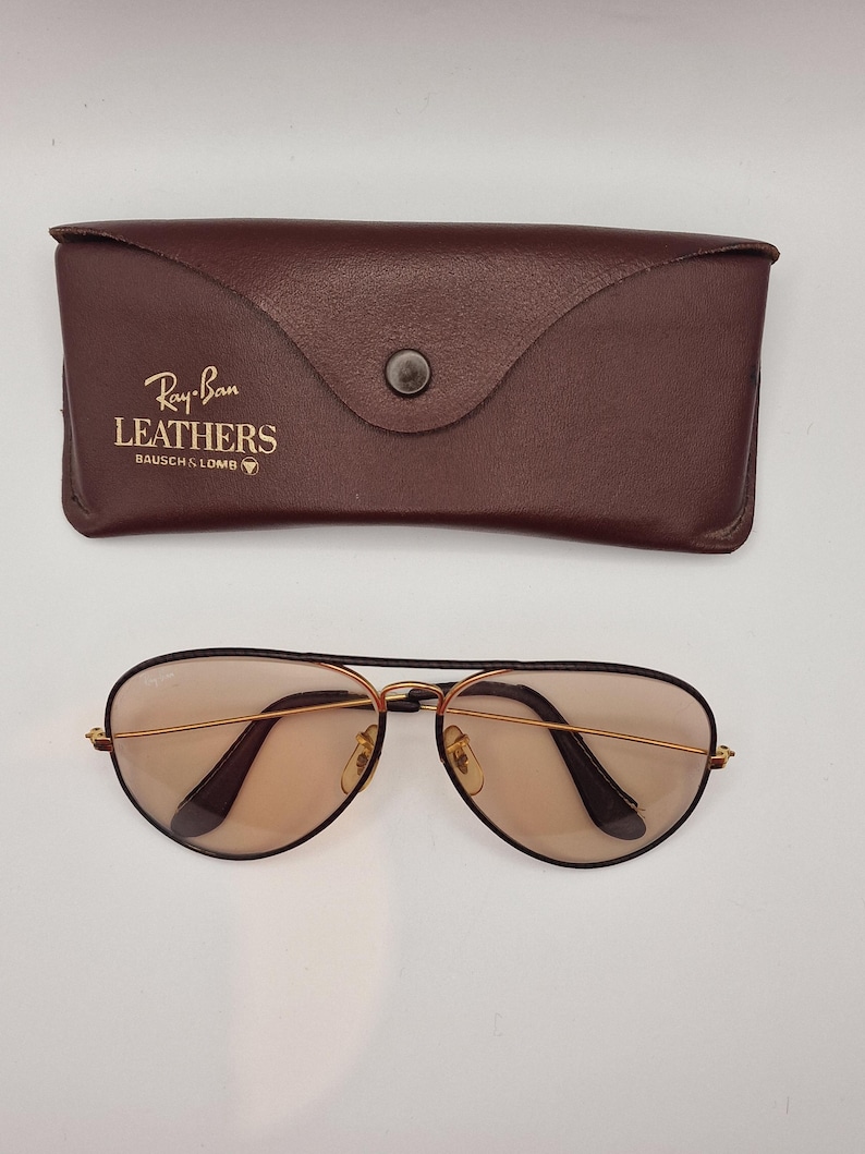 K&ouml;nnte beinhalten: Braunes Leder-Sonnenbrillenetui mit dem Text "Ray-Ban LEATHERS BAUSCH & LOMB" und einer Pilotenbrille. Die Sonnenbrille hat goldfarbene Rahmen und braune Gl&auml;ser. Das Etui hat einen Druckknopfverschluss.