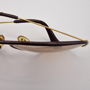 K&ouml;nnte beinhalten: Nahaufnahme einer Sonnenbrille mit braun lederummantelten Rahmen und goldfarbenen Metallakzenten. Die Gl&auml;ser sind hellbraun get&ouml;nt. Die Sonnenbrille hat einen klassischen Aviator-Stil.