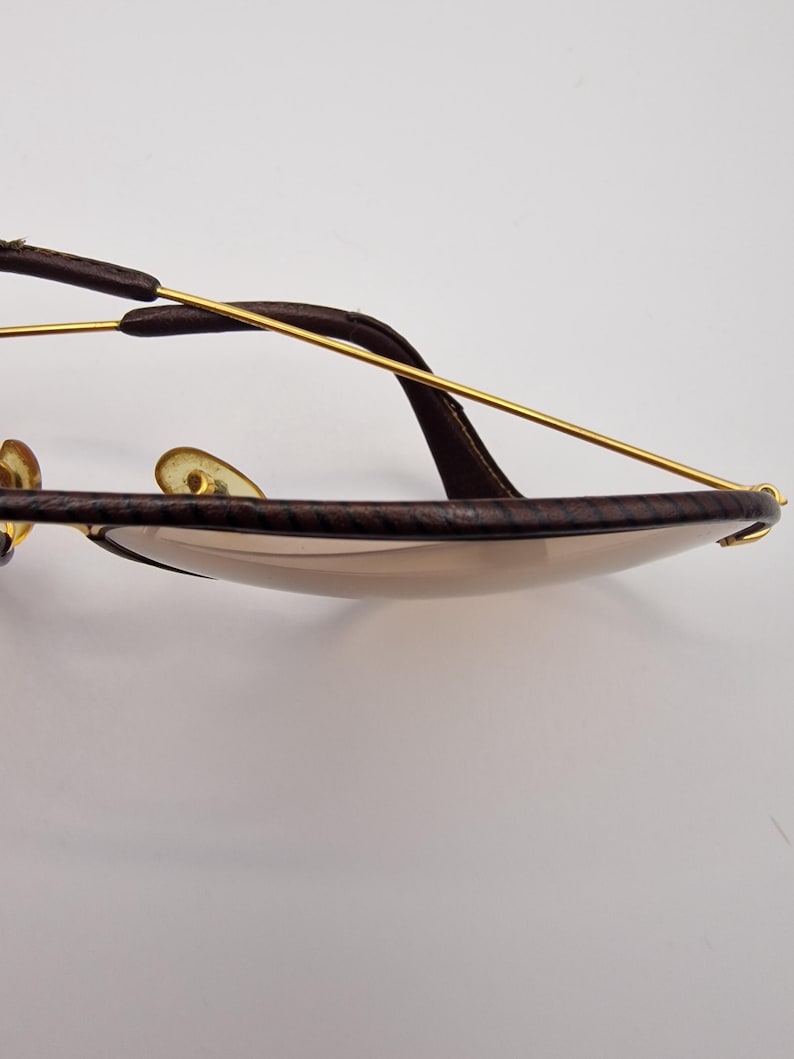 K&ouml;nnte beinhalten: Nahaufnahme einer Sonnenbrille mit braun lederummantelten Rahmen und goldfarbenen Metallakzenten. Die Gl&auml;ser sind hellbraun get&ouml;nt. Die Sonnenbrille hat einen klassischen Aviator-Stil.