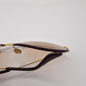 K&ouml;nnte beinhalten: Nahaufnahme einer Sonnenbrille mit braunen Gl&auml;sern und einem dunkelbraunen Lederrahmen. Die Sonnenbrille hat goldfarbene Metallakzente und ein geschwungenes Design. Die Gl&auml;ser sind hellbraun.