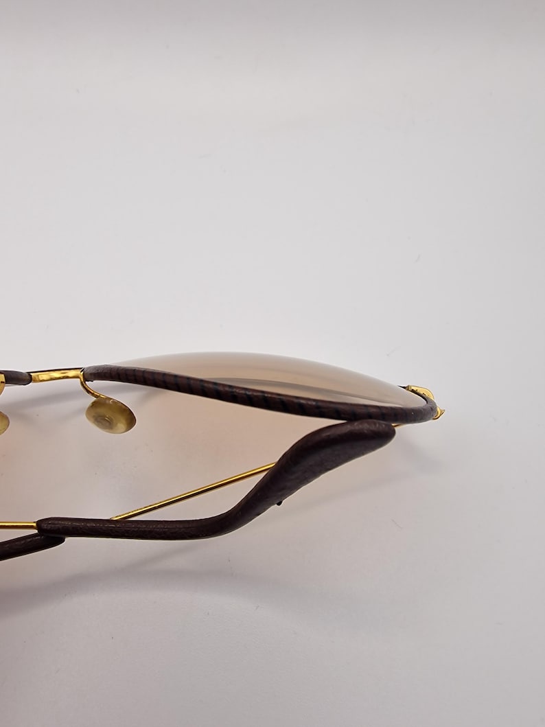 K&ouml;nnte beinhalten: Nahaufnahme einer Sonnenbrille mit braunen Gl&auml;sern und einem dunkelbraunen Lederrahmen. Die Sonnenbrille hat goldfarbene Metallakzente und ein geschwungenes Design. Die Gl&auml;ser sind hellbraun.