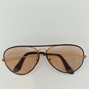 K&ouml;nnte beinhalten: Aviator-Sonnenbrille mit goldfarbenem Metallrahmen und braunen Gl&auml;sern. Die Sonnenbrille hat eine klassische Tropfenform und dunkelbraune Akzente an den B&uuml;geln und am oberen Rahmen. Die Gl&auml;ser sind hellbraun.