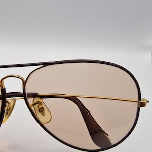 K&ouml;nnte beinhalten: Sonnenbrille im Aviator-Stil mit einem goldfarbenen Metallrahmen und braunen Gl&auml;sern. Die Gl&auml;ser haben eine Tropfenform, und der Rahmen hat einen Doppelsteg. Die B&uuml;gel sind dunkelbraun. Auf wei&szlig;em Hintergrund.