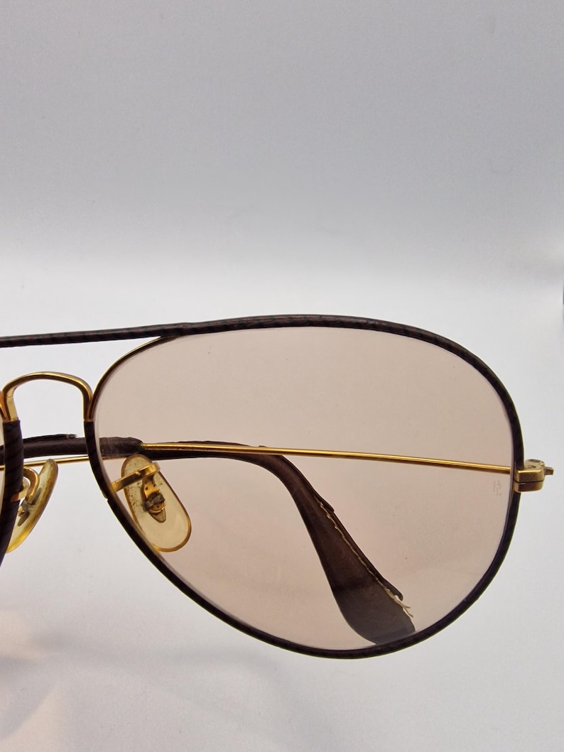 K&ouml;nnte beinhalten: Sonnenbrille im Aviator-Stil mit einem goldfarbenen Metallrahmen und braunen Gl&auml;sern. Die Gl&auml;ser haben eine Tropfenform, und der Rahmen hat einen Doppelsteg. Die B&uuml;gel sind dunkelbraun. Auf wei&szlig;em Hintergrund.