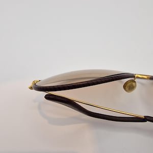 K&ouml;nnte beinhalten: Nahaufnahme einer Sonnenbrille mit braunen Gl&auml;sern und dunkelbraunen Rahmen. Die Rahmen haben goldfarbene Akzente und ein schlankes, modernes Design. Die Gl&auml;ser sind hellbraun.
