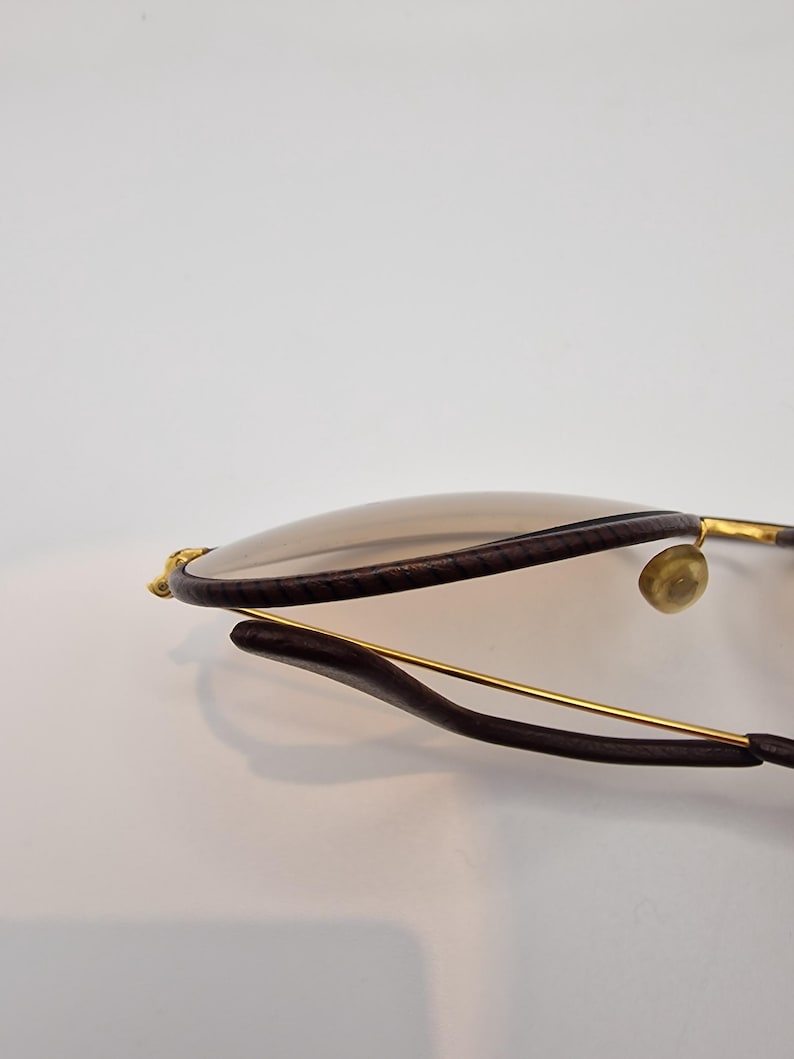 K&ouml;nnte beinhalten: Nahaufnahme einer Sonnenbrille mit braunen Gl&auml;sern und dunkelbraunen Rahmen. Die Rahmen haben goldfarbene Akzente und ein schlankes, modernes Design. Die Gl&auml;ser sind hellbraun.