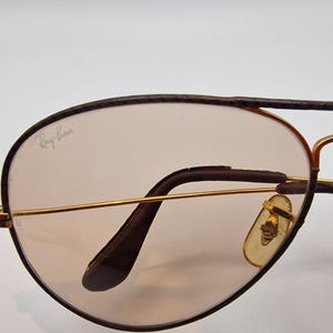 K&ouml;nnte beinhalten: Nahaufnahme einer Ray-Ban Aviator-Sonnenbrille. Die Gl&auml;ser sind hellbraun get&ouml;nt, umrahmt von goldfarbenem Metall und dunkelbraunem Besatz. Das Ray-Ban-Logo ist auf dem Glas sichtbar. Die Nasenpads sind hellgelb.