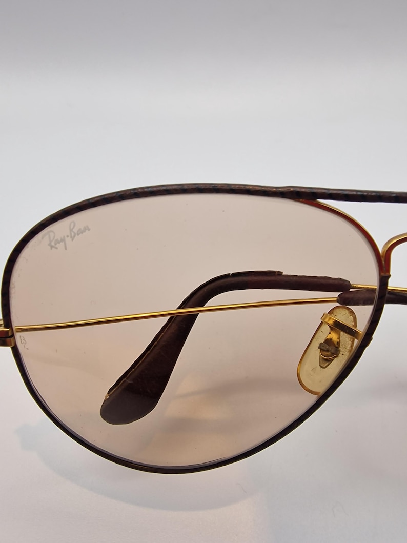K&ouml;nnte beinhalten: Nahaufnahme einer Ray-Ban Aviator-Sonnenbrille. Die Gl&auml;ser sind hellbraun get&ouml;nt, umrahmt von goldfarbenem Metall und dunkelbraunem Besatz. Das Ray-Ban-Logo ist auf dem Glas sichtbar. Die Nasenpads sind hellgelb.