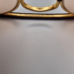 K&ouml;nnte beinhalten: Nahaufnahme einer goldgerahmten Brille mit schwarzen B&uuml;geln. Der Text "B&L RAY-BAN 62 14" ist auf dem Rahmen eingraviert. Die Gl&auml;ser sind hellrosa. Die Brille ist vor einem hellgrauen Hintergrund platziert.