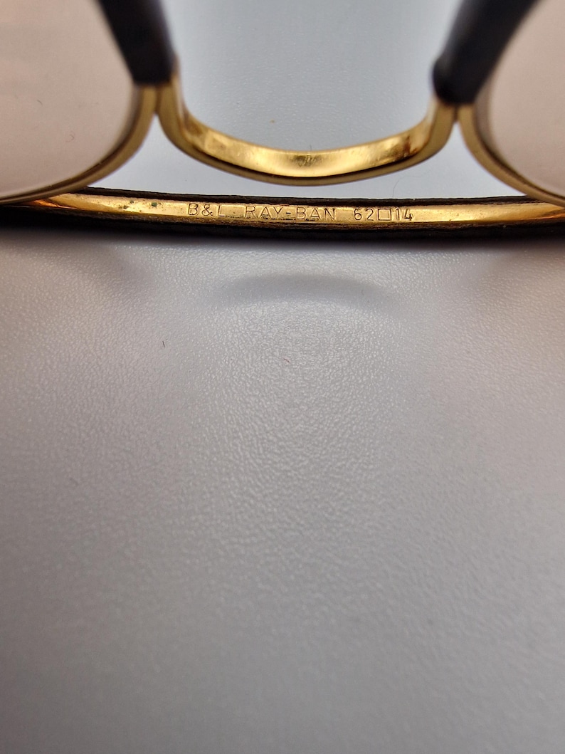 K&ouml;nnte beinhalten: Nahaufnahme einer goldgerahmten Brille mit schwarzen B&uuml;geln. Der Text "B&L RAY-BAN 62 14" ist auf dem Rahmen eingraviert. Die Gl&auml;ser sind hellrosa. Die Brille ist vor einem hellgrauen Hintergrund platziert.