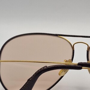 K&ouml;nnte beinhalten: Sonnenbrille im Aviator-Stil mit einem goldfarbenen Metallrahmen und braunen Gl&auml;sern. Die B&uuml;gel sind dunkelbraun mit lederartiger Textur. Die Gl&auml;ser sind hellbraun.