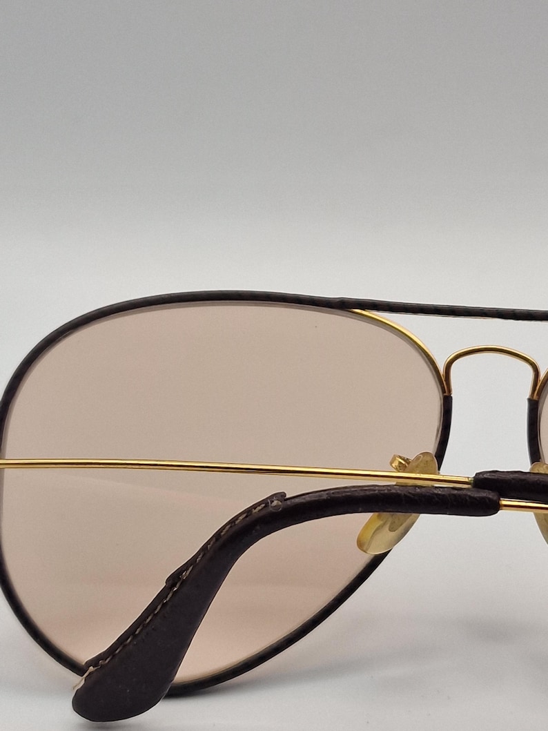 K&ouml;nnte beinhalten: Sonnenbrille im Aviator-Stil mit einem goldfarbenen Metallrahmen und braunen Gl&auml;sern. Die B&uuml;gel sind dunkelbraun mit lederartiger Textur. Die Gl&auml;ser sind hellbraun.