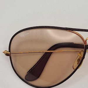 K&ouml;nnte beinhalten: Nahaufnahme einer Ray-Ban Sonnenbrille. Die Sonnenbrille hat einen goldfarbenen Rahmen mit schwarzem oberem Rand und B&uuml;geln. Die Gl&auml;ser sind hellbraun. Das Ray-Ban-Logo ist auf dem Glas sichtbar.