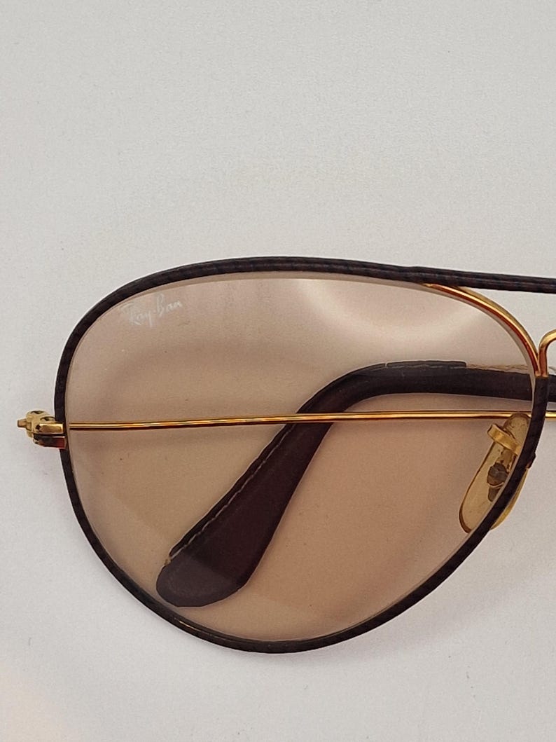 K&ouml;nnte beinhalten: Nahaufnahme einer Ray-Ban Sonnenbrille. Die Sonnenbrille hat einen goldfarbenen Rahmen mit schwarzem oberem Rand und B&uuml;geln. Die Gl&auml;ser sind hellbraun. Das Ray-Ban-Logo ist auf dem Glas sichtbar.