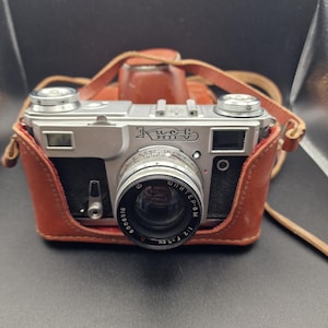 Puede incluir: Una cámara vintage plateada y negra con una lente y el texto "Kiev" en la parte delantera. La cámara está en una funda de cuero marrón con una correa a juego. Tiene un diseño retro.