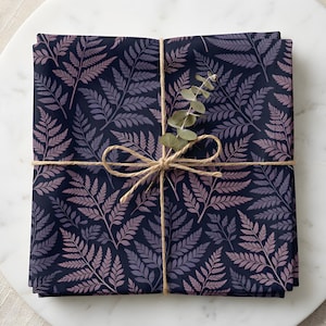 May include: A folded fabric with a dark navy background and a repeating pattern of fern leaves in shades of lavender and dusty rose. The fabric is tied with twine and adorned with a sprig of eucalyptus.