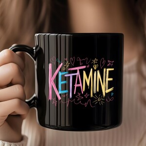 May include: Black ceramic mug with a white handle. The mug has a colorful design with the word "KETAMINE" written in pink, blue, yellow, and purple. The design also includes hearts, stars, and other doodles.