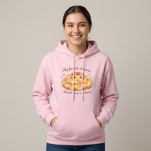 May include: A light pink hooded sweatshirt featuring a graphic of a coiled yellow snake wearing a flower crown. The text above and below the snake reads, My favorite banana doesn't grow on trees, accompanied by two small pink hearts on each side.