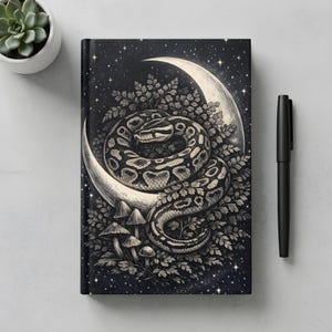 May include: A rectangular notebook featuring a black and white illustration of a coiled snake resting on a crescent moon surrounded by ferns and mushrooms against a starry night sky, accompanied by a black pen and a small potted succulent plant.