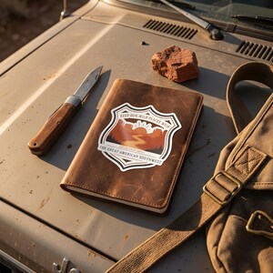 Puede incluir: Un cuaderno de cuero marrón con una pegatina en forma de escudo que muestra un paisaje desértico, el texto "KEEP OUR WILD PLACES WILD" y "THE GREAT AMERICAN SOUTHWEST," una navaja plegable con mango de madera y una piedra roja.