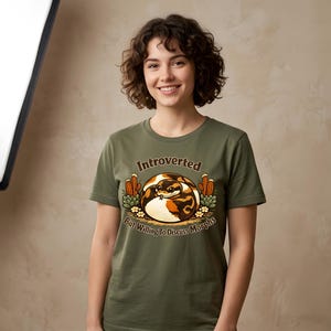 May include: An olive green t-shirt featuring a graphic of a coiled ball python surrounded by cacti and small flowers. The text above the snake reads "Introverted" and the text below reads "But Willing To Discuss Morphs" in brown lettering.