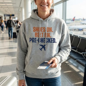 Könnte beinhalten: Ein grauer Kapuzenpullover mit dem Text "SHOES ON. BELT ON. PRE-CHECKED." in Orange und Marineblau. Eine marineblaue Flugzeuggrafik befindet sich unter dem Text. Die Person hält einen Reisepass und eine Bordkarte.