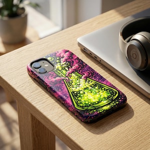 May include: A phone case featuring a neon green and yellow beaker design with pink and purple smoke. The case is on a wooden table next to a laptop and headphones.
