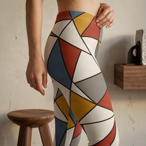 May include: Leggings featuring a geometric pattern of triangles in shades of cream, muted blue, mustard yellow, and terracotta red, separated by bold black lines. The design covers the entire surface of the fitted garment, creating a structured look.