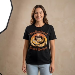 Może przedstawiać: Czarny t-shirt z krótkim rękawem z grafiką przedstawiającą żółto-szarego węża owiniętego wokół czarnego kubka do kawy. Napis "Professional Snoot Booper" jest nadrukowany zakrzywioną brązową czcionką nad i pod okrągłym wzorem zachodzącego słońca.