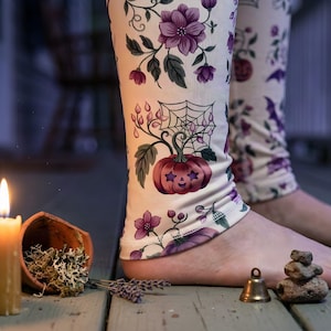 May include: Cream-colored leggings featuring a repeating pattern of purple flowers, green leaves, spiderwebs, and orange pumpkins with star-shaped eyes. The fabric has a fitted silhouette that covers the lower legs and ankles with a hemmed finish.