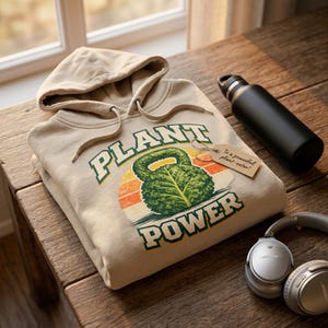 May include: A tan hoodie with the words "PLANT POWER" and a graphic of a leafy kettlebell. A black water bottle and silver headphones are next to the hoodie on a wooden surface. A small tag reads "To a powerful plant-eater!"