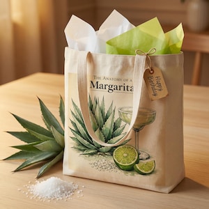 May include: A beige canvas tote bag featuring the words "The Anatomy of a Margarita" and a watercolour illustration of an agave plant, margarita glass, and lime. The bag has a "Happy Birthday" tag and is filled with white and green tissue paper.