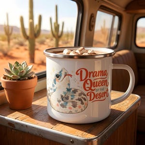 May include: A white enamel mug filled with a hot beverage topped with marshmallows. The mug features a graphic of a coiled snake with blue and grey scales and the red text "Drama Queen of the Desert" with small star accents printed on the side.