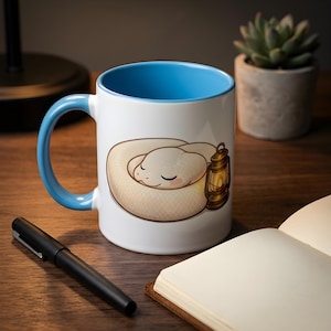 May include: A white ceramic mug with a bright blue interior and handle. The exterior features a graphic illustration of a small, cream-colored snake coiled in a circle next to a lit, vintage-style brass lantern with a warm, glowing flame.