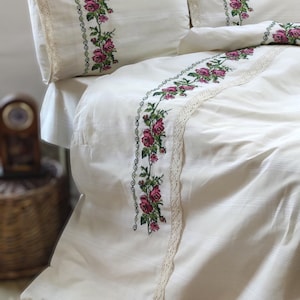 May include: White bedding with a delicate lace trim and embroidered floral design featuring pink roses and green leaves.
