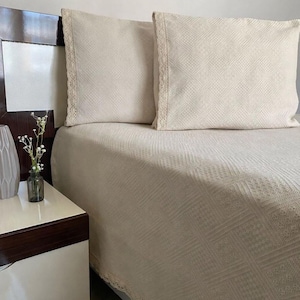May include: Cream-colored bedding set featuring two square pillows and a matching bedspread. The pillows and bedspread have a textured, geometric pattern and are trimmed with delicate lace. A nightstand with a vase of flowers is visible.